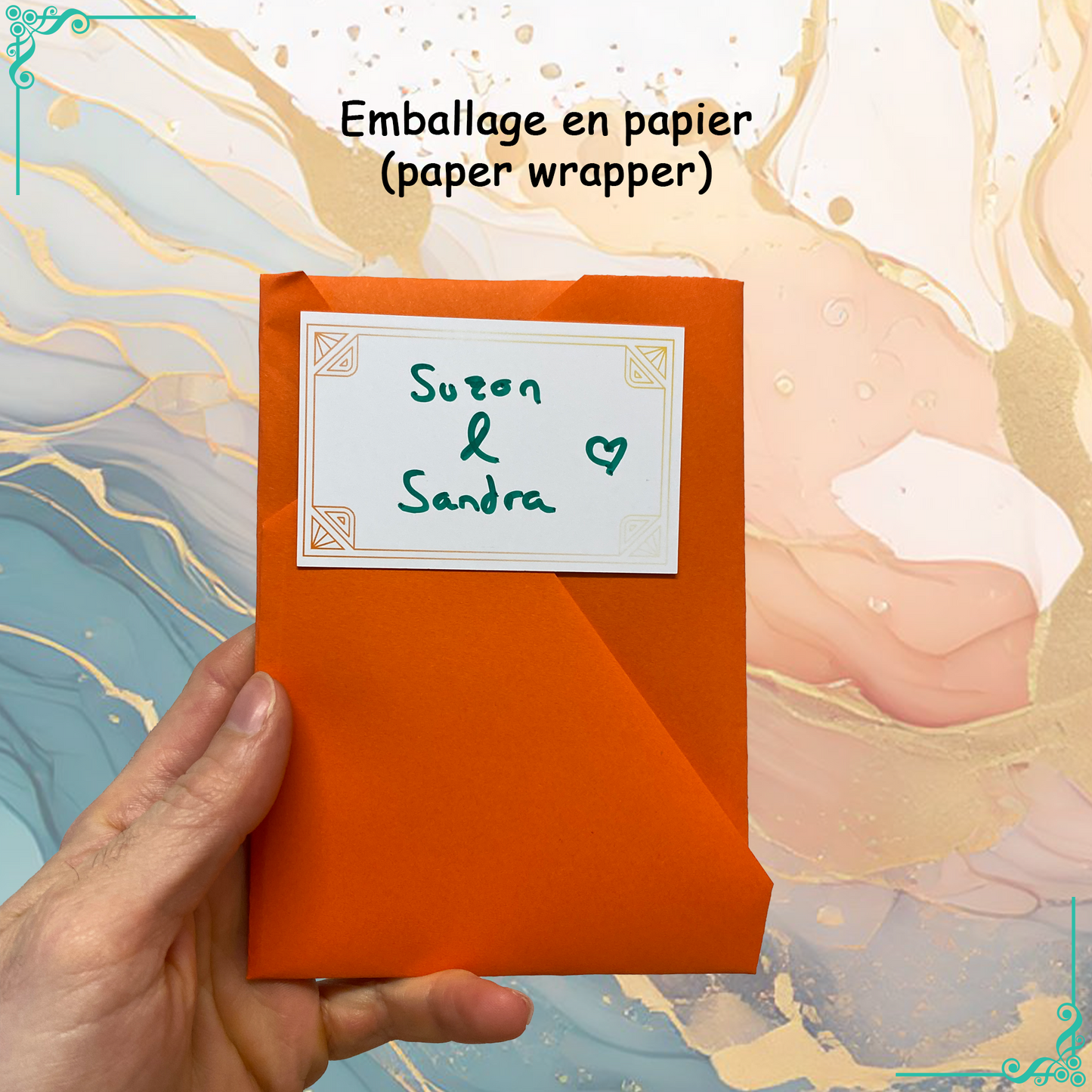 Emballage en papier orange contenant un aimant personnalisé. Étiquette manuscrite avec les noms "Suzon & Sandra" et un cœur dessiné. Texte au-dessus : "Emballage en papier (paper wrapper)". Présentation élégante et personnalisée.