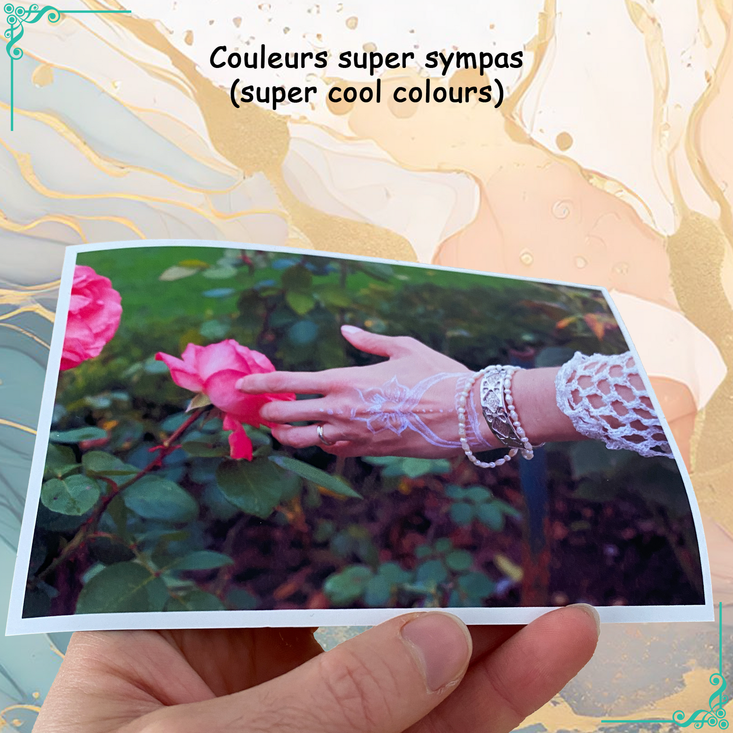 Photo imprimée avec des couleurs vives et éclatantes. L'image montre une main ornée de bracelets touchant une rose dans un jardin fleuri. Texte au-dessus : "Couleurs super sympas".