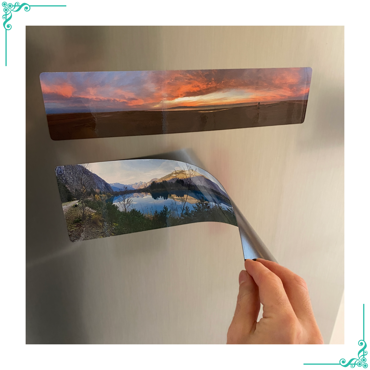 Main tenant un aimant panoramique flexible imprimé avec un paysage de lac entouré de montagnes, montrant l'épaisseur fine et la flexibilité de l'aimant. Un autre aimant représentant un coucher de soleil est fixé sur le réfrigérateur.