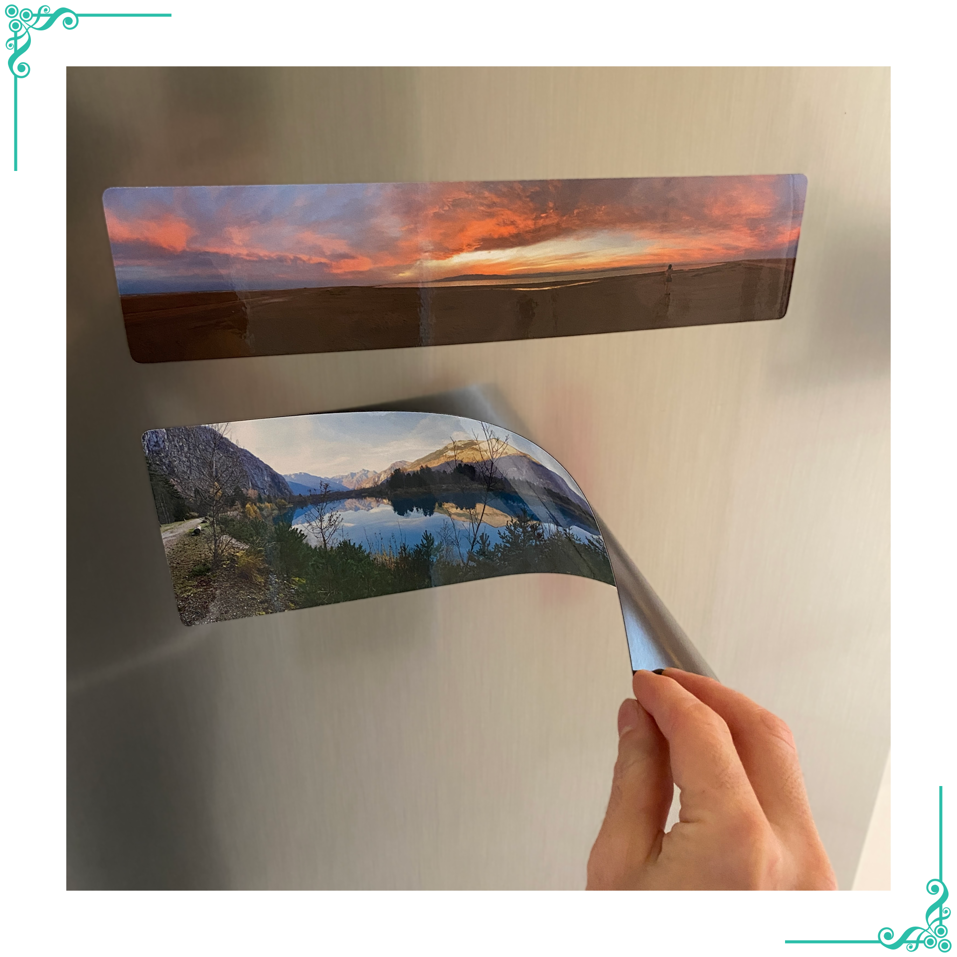 Main tenant un aimant panoramique flexible imprimé avec un paysage de lac entouré de montagnes, montrant l'épaisseur fine et la flexibilité de l'aimant. Un autre aimant représentant un coucher de soleil est fixé sur le réfrigérateur.