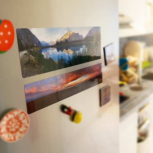 Deux aimants panoramiques fixés sur un réfrigérateur. Les images imprimées montrent des paysages naturels : un lac entouré de montagnes et un coucher de soleil sur une plage.